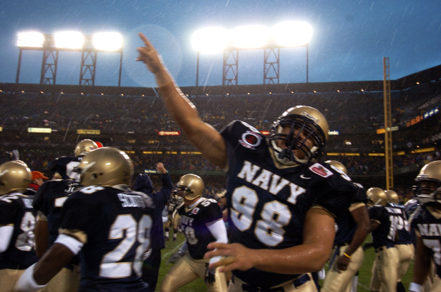 Navy-Fußballspieler in Helmen und Uniformen feiern ein Touchdown auf einem regennassen Rasenfeld, mit Zuschauern und Stadionbeleuchtung im Hintergrund.