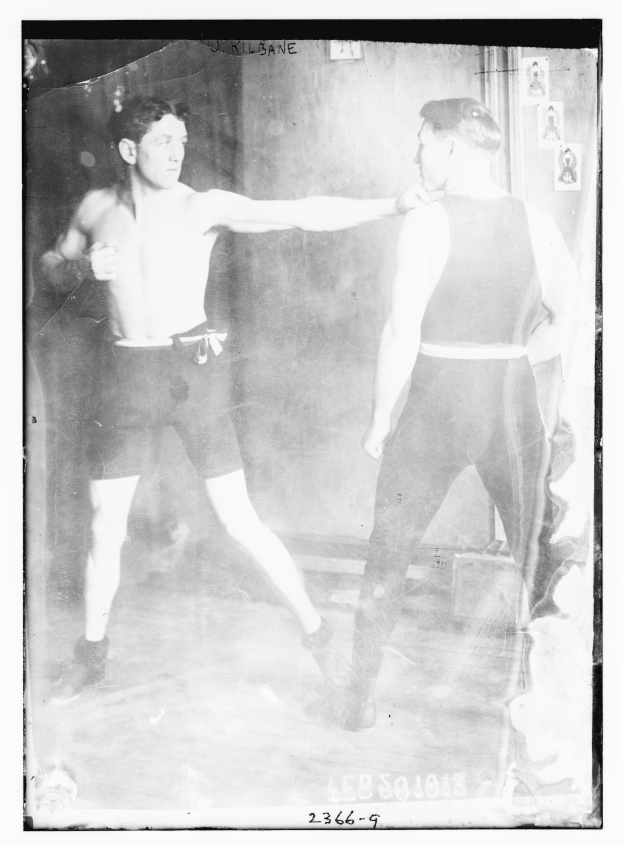 Schwarz-weiß-Foto von zwei Boxern im Ring mit Plakaten an der Hintergrundwand und Text unten.