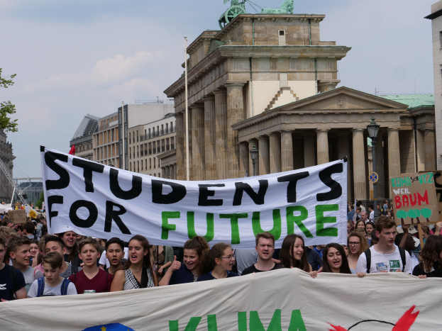 Gruppe von Schülern marschiert in Berlin mit buntem "Students for Future"-Schild vor Gebäuden, Bäumen und Himmel.