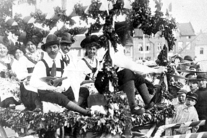 Schwarzes und weißes Bild eines Paradewagens mit Menschen in traditioneller bayrischer Tracht, einige spielen Instrumente, mit Blattdekorationen vor Buildings und einem klaren Himmel.