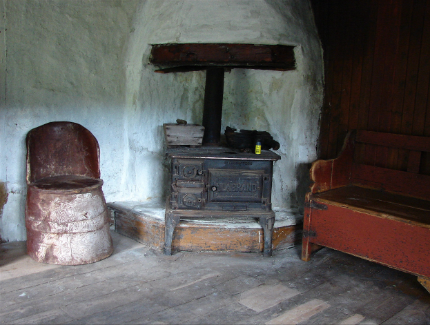 Altmodischer Holzofen in einem Raum mit einer hölzernen Bank auf der rechten Seite, einem Stuhl auf der linken Seite und einer hölzernen Wand im Hintergrund.
