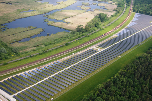 Luftaufnahme einer Solar-Farm mit Solar-Panelen auf einem Feld, umgeben von Bäumen, Gras, Pflanzen und Wasser, mit einem Zug auf einer Bahnstrecke.