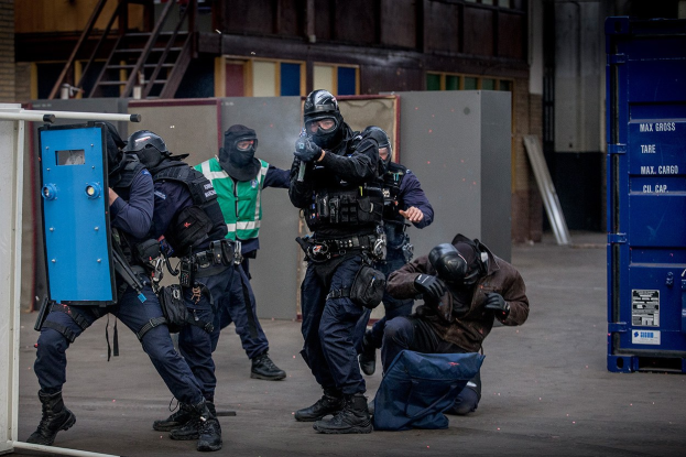Gruppe von Polizisten in Einsatzausrüstung vor einem Gebäude stehend, mit einer Person im Vordergrund am Boden liegend.