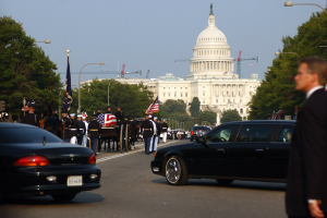 Trauerzug mit Menschen in Mützen und Fahnen vor dem Kapitolsgebäude, Fahrzeuge auf der Straße, Laternenpfähle, Bäume und Gebäude unter einem klaren Himmel.
