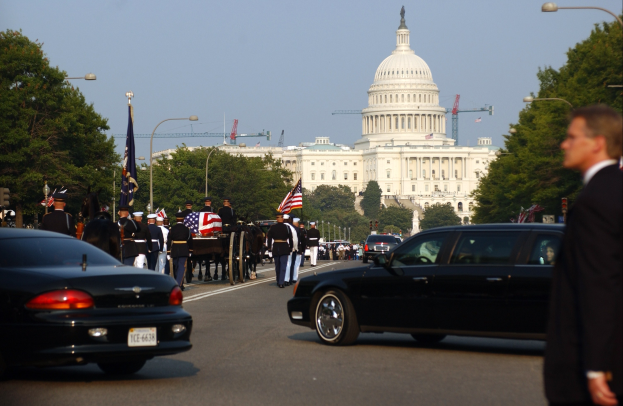 Trauerzug mit Menschen in Mützen und Fahnen vor dem Kapitolsgebäude, Fahrzeuge auf der Straße, Laternenpfähle, Bäume und Gebäude unter einem klaren Himmel.