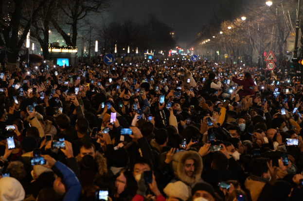 Eine große Menschenmenge, die nachts auf einer Straße steht, beleuchtet von Straßenlaternen und umgeben von Bäumen und Schildern, viele halten ihre Handys hoch.