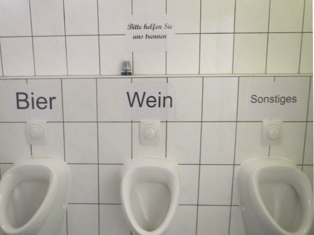 Drei weiße Toiletten an einer Wand montiert mit Schildern darüber, die 'Bier Wein' und 'Sonstiges' lesen.