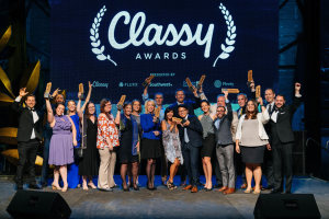 Gruppe von Menschen auf einer Bühne, die Auszeichnungen halten, mit Rednern darunter und einem Bildschirm im Hintergrund mit 'Classy Awards 2019'