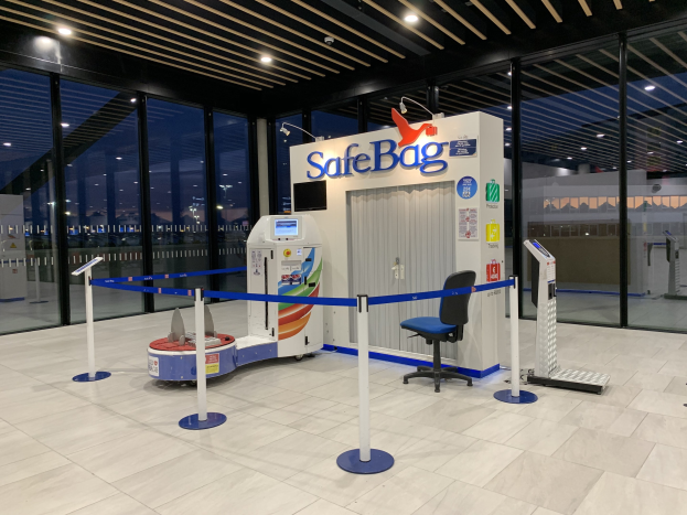 Sicherheitskontrolle am Flughafen mit Stuhl, Absperrgittern, Maschinen, Bildschirm, bedrucktem Schild, Glaswänden, Deckenleuchten und sichtbarem Boden.
