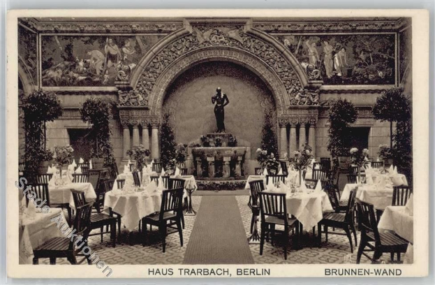 Schwarzes und weißes Foto eines alten Berliner Restaurantinnenraums mit Tischen, die mit Vasen gedeckt sind, Stühlen, einer Statue im Hintergrund, geschnitzten Wänden, Pflanzen und Text unten.