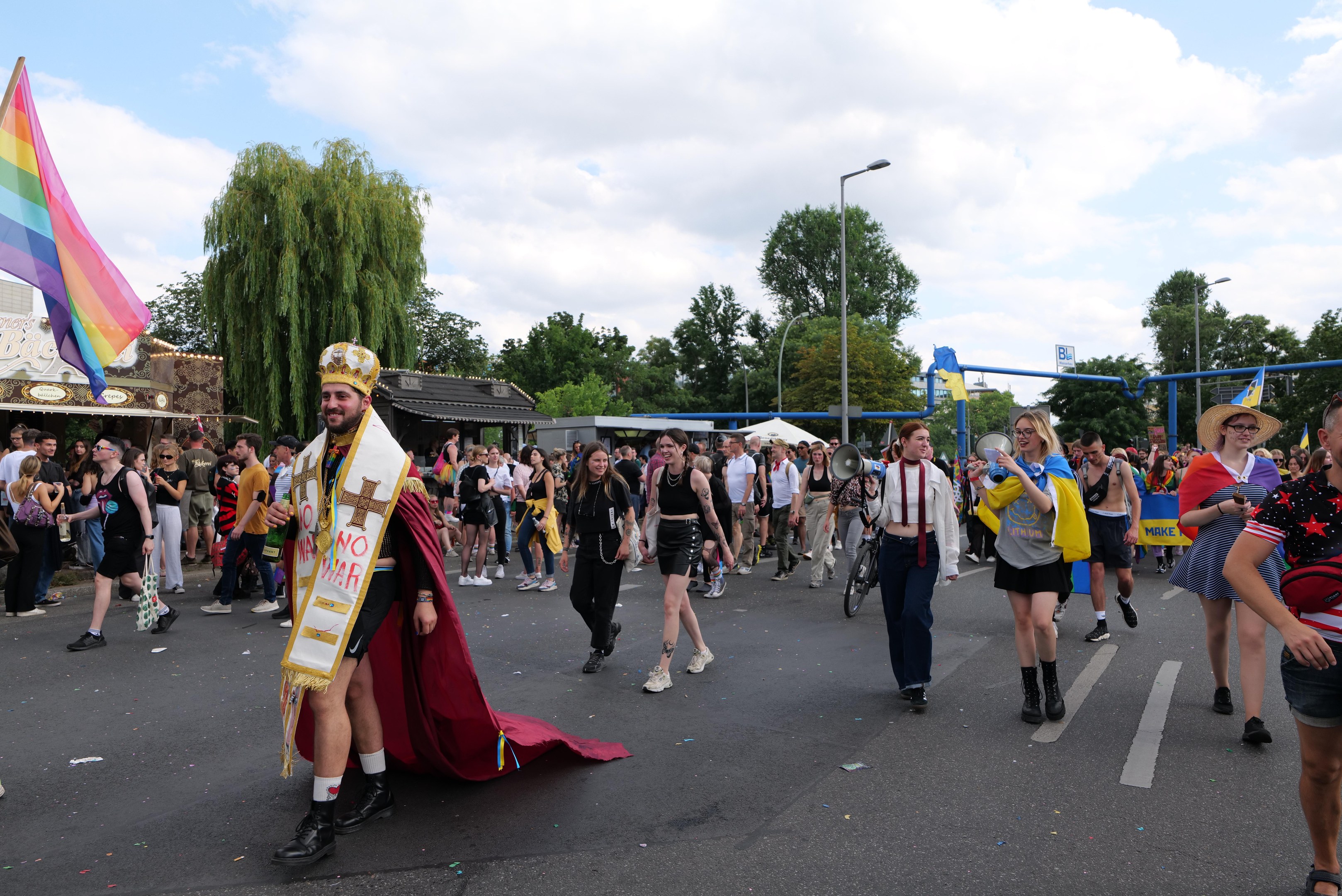 Menschen marschieren in einem Umzug mit einer Regenbogenfahne und Musikinstrumenten, mit Laternenmasten, Bäumen, Hütten und Wolken im Hintergrund.