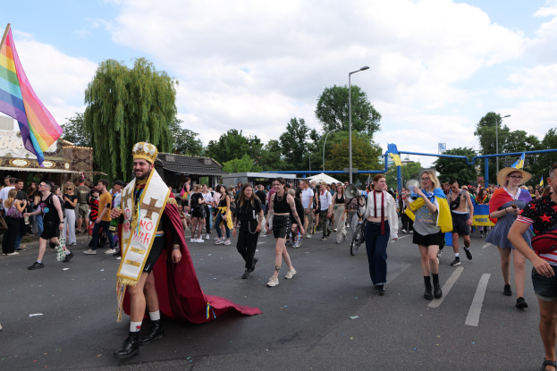 Menschen marschieren in einem Umzug mit einer Regenbogenfahne und Musikinstrumenten, mit Laternenmasten, Bäumen, Hütten und Wolken im Hintergrund.
