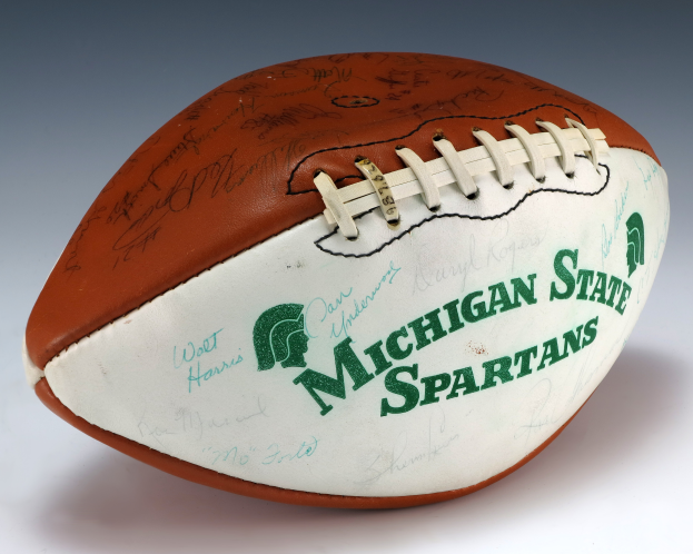 Eine weiße Fläche mit einem signierten Football der Michigan State Spartans, das das Team-Logo und Text trägt.