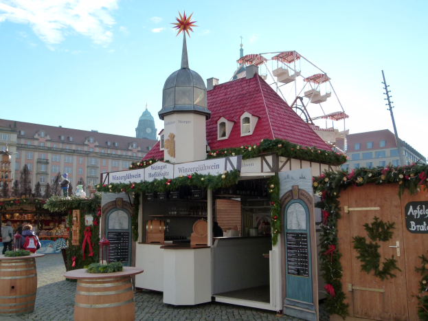 Ein kleines Gebäude mit rotem Dach und einem Riesenrad davor, umgeben von Menschen und Festobjekten, mit Gebäuden, Bäumen und einem bewölkten Himmel im Hintergrund auf einem Oktoberfest-Event in München, Deutschland.