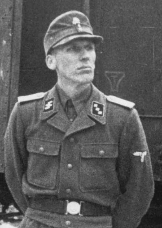 Ein Schwarz-Weiß-Foto eines Mannes in Uniform, der eine Mütze trägt und vor einem Zug steht, mit Text unten, der besagt, dass er mutmaßlich ein deutscher Soldat ist.