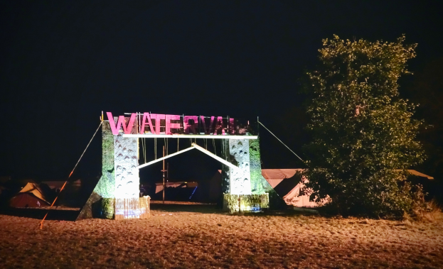 Bühnenaufbau für ein nächtliches Musikfestival mit einem Bogen mit Text, einem Baum auf der rechten Seite, Zelten im Hintergrund und einem dunklen Himmel.