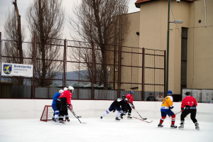 Menschen, die Eis-hockey auf einer Eisbahn mit Geb√§uden, B√§umen, einer Stra√Ÿenlaterne, einer Namensplakette und Z√§unen im Hintergrund unter einem Himmel spielen.