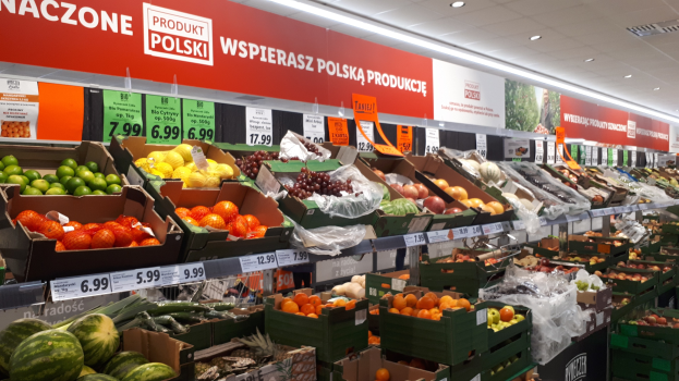 Ein Geschäft mit verschiedenen Obst- und Gemüsesorten in Kartons, jeweils mit Preisschildern, und informativen Schildern an den Wänden unter Deckenlampen.