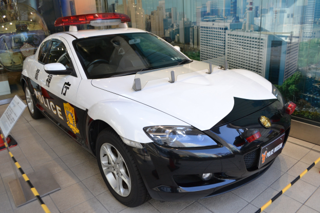 Polizeiauto vor einer Glaswand geparkt, mit einem Rad sichtbar, auf einem Boden neben einer Texttafel; Gebäude, Bäume und Himmel durch die Glaswand sichtbar.