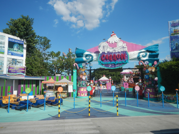 Karussell am Parkeingang mit Straße darunter, eingezäunter Bereich, Buden, Banner, Bäume und bewölkter Himmel im Hintergrund.