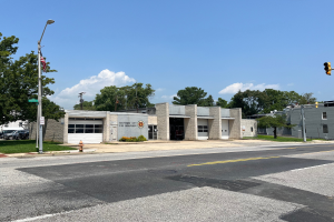 Eine Feuerwache an einer Straßenecke mit umliegenden Gebäuden, Fahrzeugen, Verkehrszeichen, einer Hydranten, Grünflächen und einem bewölkten Himmel.