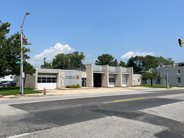 Eine Feuerwache an einer Straßenecke mit umliegenden Gebäuden, Fahrzeugen, Verkehrszeichen, einer Hydranten, Grünflächen und einem bewölkten Himmel.