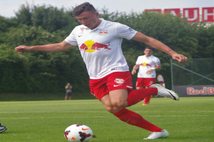 Ein professioneller Fußballspieler kickt einen Ball auf einem grünen Feld mit Bäumen und einem klaren blauen Himmel im Hintergrund, in der Nähe eines Schildes, auf dem "RB Leipzig" steht.