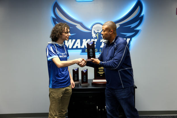 Zwei Männer schütteln sich vor einer mit einem Logo versehenen Wand die Hand, einer hält eine Trophäe, während Auszeichnungen auf einem nahen Tisch liegen.