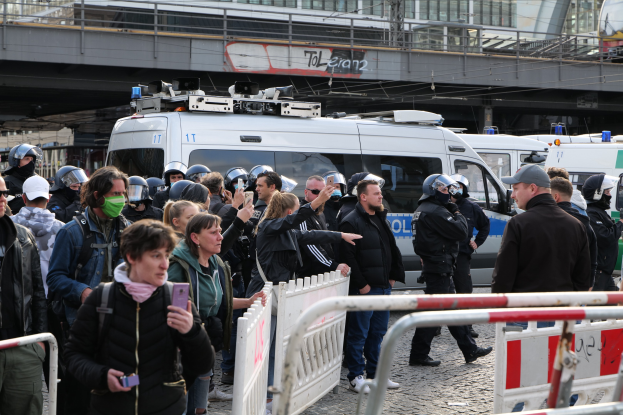 Eine Gruppe von Menschen, einige mit Helmen und Handys, steht vor Polizeifahrzeugen mit Barrieren im Vordergrund, eine Brücke und Gebäude im Hintergrund während einer Demonstration in Berlin.