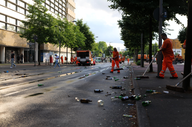 Menschen in orangen Uniformen sammeln Müll von einer Straße mit Flaschen und Schutt, mit Bäumen, Pfählen und Fahrzeugen im Hintergrund.