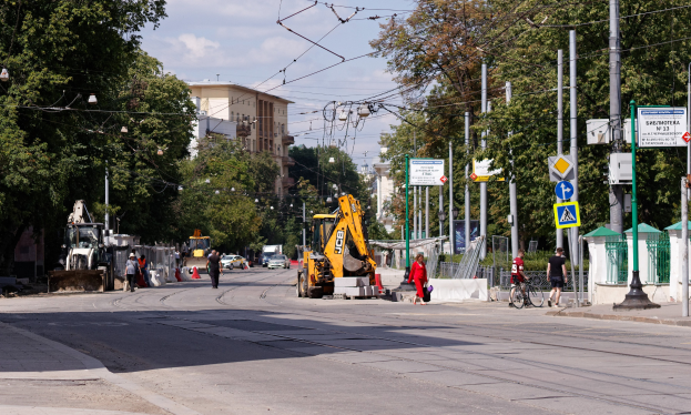 Stadtstraße mit Baustelle, Fahrzeugen, Füßgängern, einem Radfahrer, Verkehrskegeln, Masten, Schildern, Strommästen mit Drähten, Bäumen, Gebäuden mit Fenstern und einem bewölkten Himmel.
