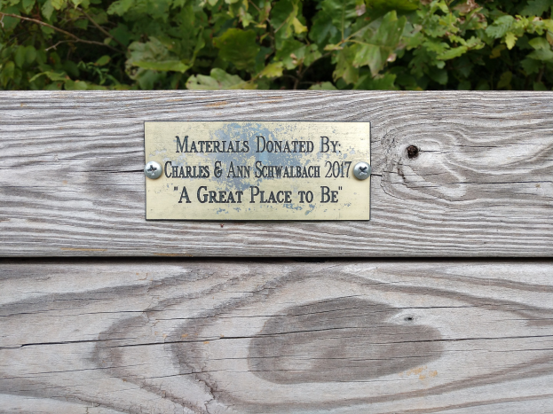 Holzbank mit einer Tafel, auf der "Materialien gespendet von Charles & Ann Schwabach 2017: Ein toller Ort" steht, in einem Garten mit üppiger grüner Vegetation.