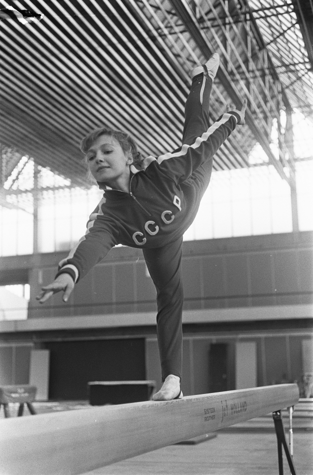 Ein junges Mädchen führt Gymnastik auf einem Balancebalken in einer Turnhalle durch, mit einer Wand im Hintergrund und einer Decke mit Stangen oben.
