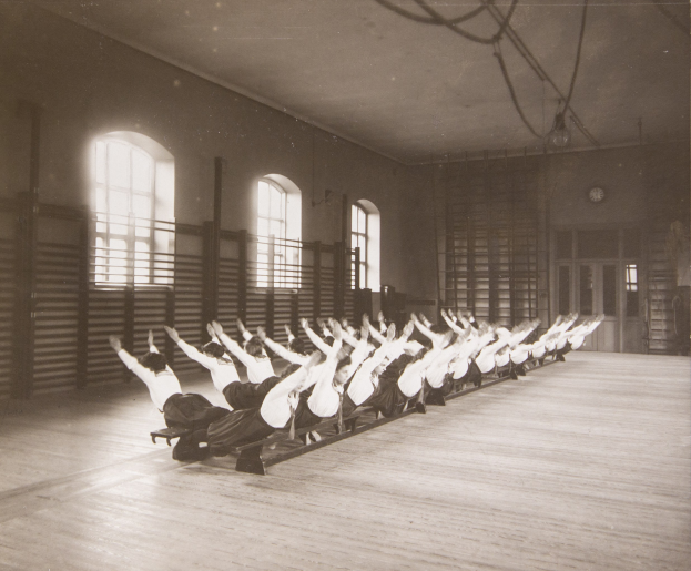 Gruppe von Menschen, die Pilates-Übungen in einem Fitnessstudio mit einer Uhr an der Wand durchführen.