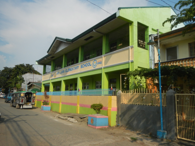 Ein Schulgebäude mit einer grünen und gelben Fassade neben einer Straße, das Fahrzeuge, Topfpflanzen, einen Zaun, ein Tor, einen Straßenpfahl, Bäume, Drähte und einen bewölkten Himmel zeigt.