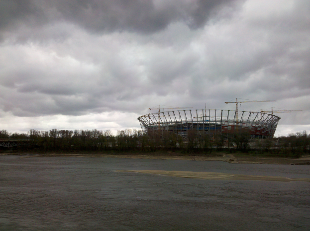 Das Olympiastadion in Kiew, Ukraine, ein großes Stadion umgeben von Bäumen und einer Brücke, mit einem bewölkten Himmel im Hintergrund und sichtbarem Boden unten.
