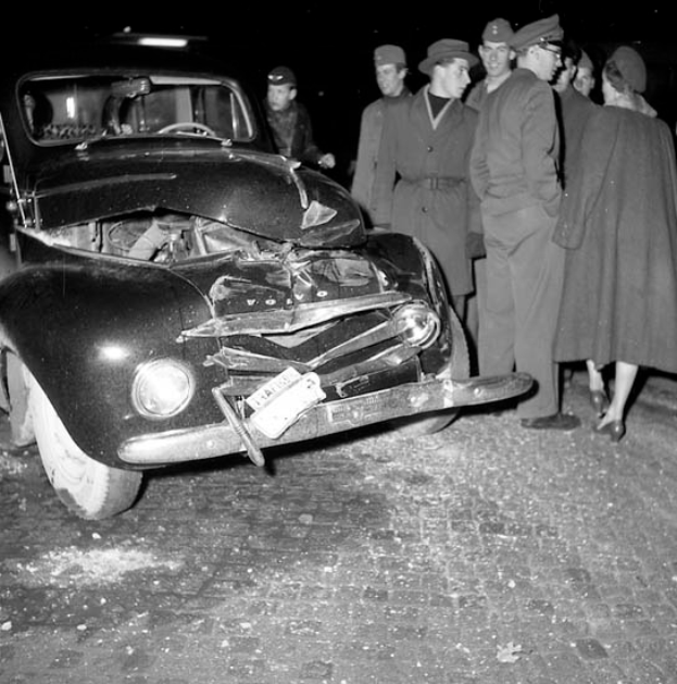 Eine Schwarz-Weiß-Szene eines beschädigten Autos am Straßenrand, umgeben von einer Gruppe von Menschen, die besorgt auf das Auto schauen.