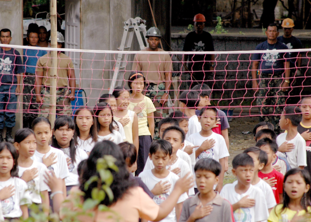 Gruppe von Kindern vor einem Netz mit einer Frau im Vordergrund, Hintergrund enthält Menschen in Helmen, eine Leiter, eine Wand und Bäume.