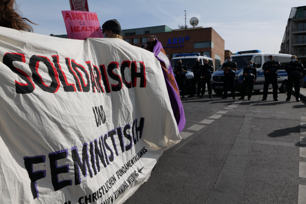 Gruppe von Menschen marschiert auf einer Straße und hält eine "Solidarität und Feminismus"-Schlagzeile hoch, mit geparkten Fahrzeugen und Gebäuden im Hintergrund unter einem klaren blauen Himmel.