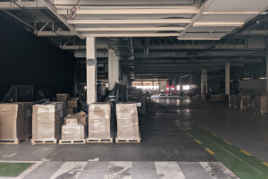 Großer Lagerraum mit zahlreichen Kartonagen auf Paletten, beleuchtet von Deckenlampen und gestützt von tragenden Säulen, mit einer Deckenanzeige, die ein Flottenmanagementsystem anzeigt.