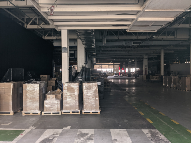 Großer Lagerraum mit zahlreichen Kartonagen auf Paletten, beleuchtet von Deckenlampen und gestützt von tragenden Säulen, mit einer Deckenanzeige, die ein Flottenmanagementsystem anzeigt.