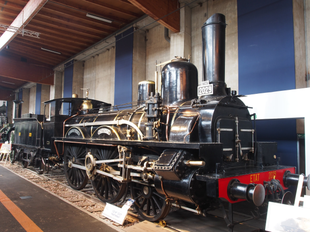 Eine gro├če, alte Dampflokomotive in einem Museum Setting umgeben von Eisenbahnschienen, Informationsschildern und anderen Ausstellungen, mit einer Wand und beleuchteter Decke im Hintergrund.