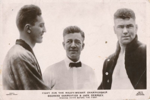 Drei Männer in einem Schwarz-Weiß-Foto, der Mittlere in einem weißen Hemd und einer Schleife flankiert von zwei Männern in Anzügen, mit Text unten lesend 'Kampf um die Schwergewichtmeisterschaft'.