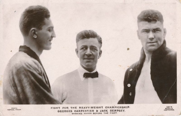Drei Männer in einem Schwarz-Weiß-Foto, der Mittlere in einem weißen Hemd und einer Schleife flankiert von zwei Männern in Anzügen, mit Text unten lesend 'Kampf um die Schwergewichtmeisterschaft'.
