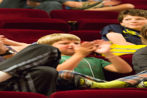 Ein Junge mit einem menschlichen Gesicht sitzt zwischen einer Gruppe von Menschen in einem Kino und klatscht in die Hände.