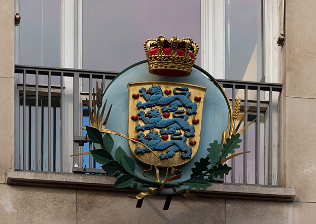 Wappen der britischen Königsfamilie an einer Gebäudewand mit einer Krone und Blätter-Logo, Fenstern und Gittern im Hintergrund.