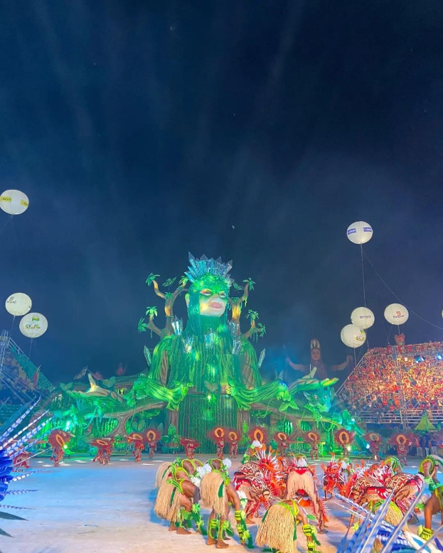 Ein lebendiger Karneval in Rio de Janeiro, Brasilien, mit Performern in bunten Kostümen auf der Bühne, einer großen Menge im Publikum und einem Himmel voller bunter Lichter und Ballons.