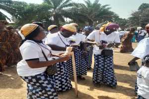 Gruppe von Frauen in weißen und schwarzen Kleidern mit einigen, die gelbe Helme tragen, auf einem staubigen Feld mit Bäumen und einem klaren blauen Himmel im Hintergrund.