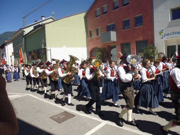 Eine Gruppe von Menschen in traditioneller bayrischer Kleidung, die auf der Straße Musikinstrumente spielen, während sie durch eine Straße mit Gebäuden gehen, einige halten Fahnen, mit einem Hügel und einem klaren blauen Himmel im Hintergrund.