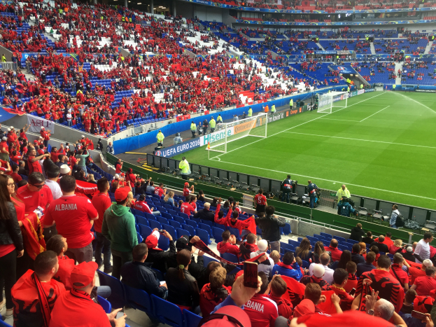 Ein volles Stadion mit Sitz- und Stehplätzen, Bannern, einem Tor und Deckenleuchten, während die Menge gespannt auf den Beginn eines Fußballspiels wartet.